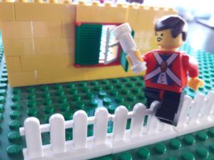 Wall of Lego house in background, Lego fence in foreground, Lego minifigure soldier holding a small club sitting on the fence