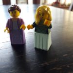 Two Lego minifigure women, one with brown hair tied back and lilac dress, one with loose blonde hair and green dress