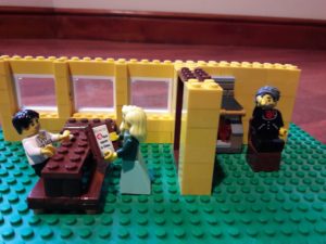 Lego room with three windows, in which a Lego minifigure girl reads a book to a Lego minifigure man. In an adjacent Lego room a Lego minifigure man listens.e